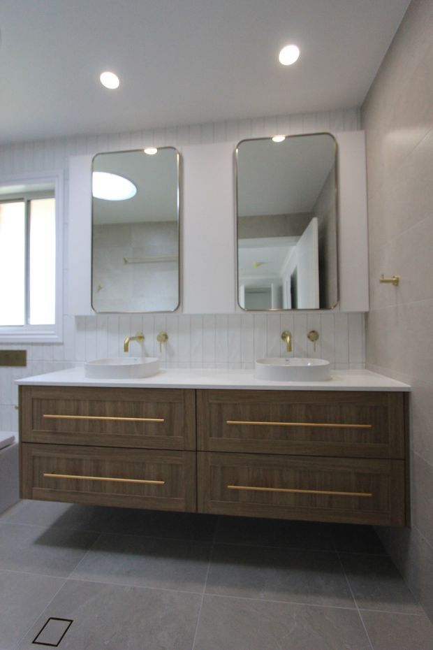 A bright contemporary bathroom featuring a wide double vanity with dark timber-look drawers, white benchtop, two round vessel basins, brushed brass taps and long handles, rounded rectangular mirrors, light wall panelling and large grey floor and wall tiles. A window and recessed ceiling lights provide natural and artificial illumination.