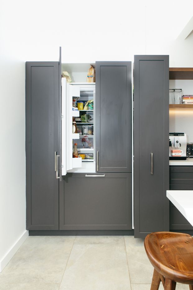A modern kitchen storage wall with tall dark grey fixed cabinetry and an open integrated refrigerator stocked with food. White walls, pale large-format tiled flooring, timber open shelving, under-shelf lighting, and a white benchtop are visible. A wooden stool appears in the foreground.