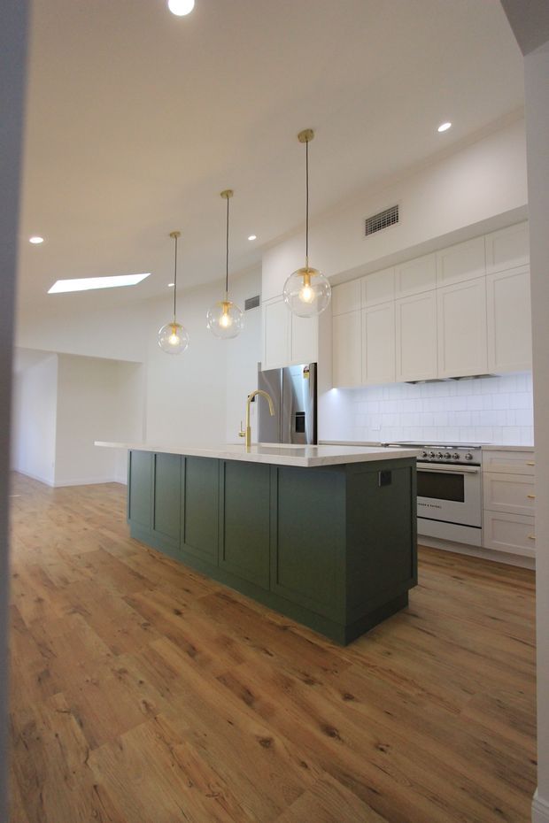 Bright contemporary kitchen viewed from the adjoining open area, featuring a large dark green island with integrated sink and brass tap, white shaker-style cabinetry, a white tiled splashback, stainless steel appliances, warm timber flooring, and three suspended globe pendant lights. A skylight and recessed ceiling lights add illumination.