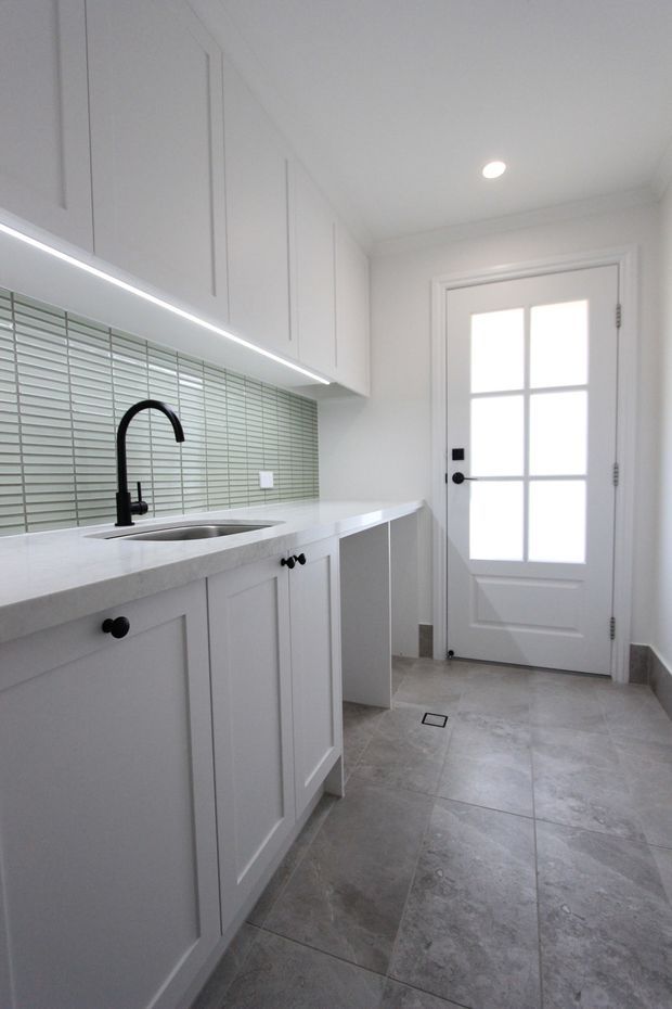 A bright, compact laundry room with white shaker-style built-in cabinetry, a white benchtop, stainless-steel sink and tall matte-black gooseneck tap. A horizontal pale green-grey tiled splashback sits beneath under-cabinet lighting, while large grey stone-look floor tiles and a white glazed-panel door complete the clean, functional space.