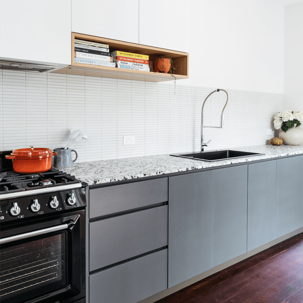 A bright contemporary kitchen with matte grey-blue flat-panel cabinetry, a white horizontally tiled splashback, and a terrazzo-style stone benchtop. The room includes a black freestanding oven and cooktop, undermount sink with a tall chrome gooseneck tap, open timber shelving with books and decor, and dark polished timber flooring.