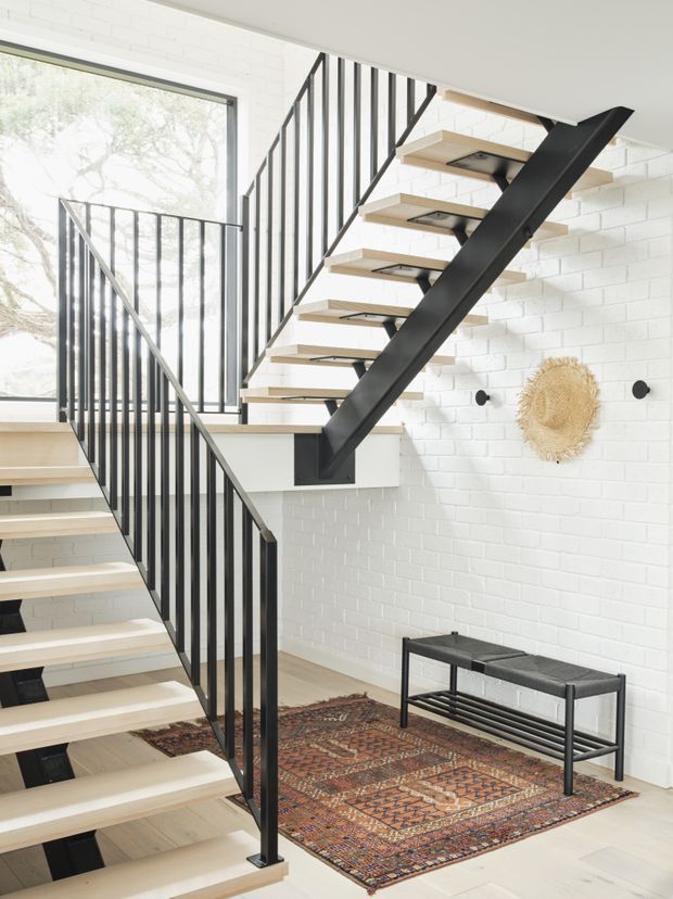 Bright contemporary interior stairway with two flights of open-riser stairs, pale timber treads, black steel stringers and slender black metal balustrades. White-painted brick walls, large glazing, light timber flooring, a patterned rug and a simple black bench create a high-contrast, minimalist-industrial look.