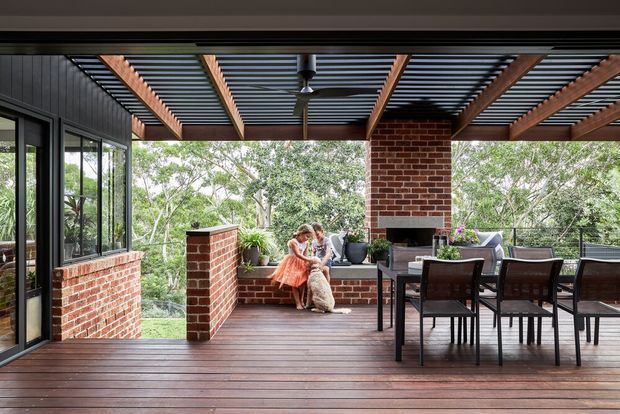 A spacious covered outdoor dining and entertaining deck with extensive glazing, dark timber decking, exposed timber pergola beams, and a corrugated translucent roof. Red brick walls and a brick outdoor fireplace or chimney feature define the space, with a large dining table and chairs overlooking leafy surroundings.