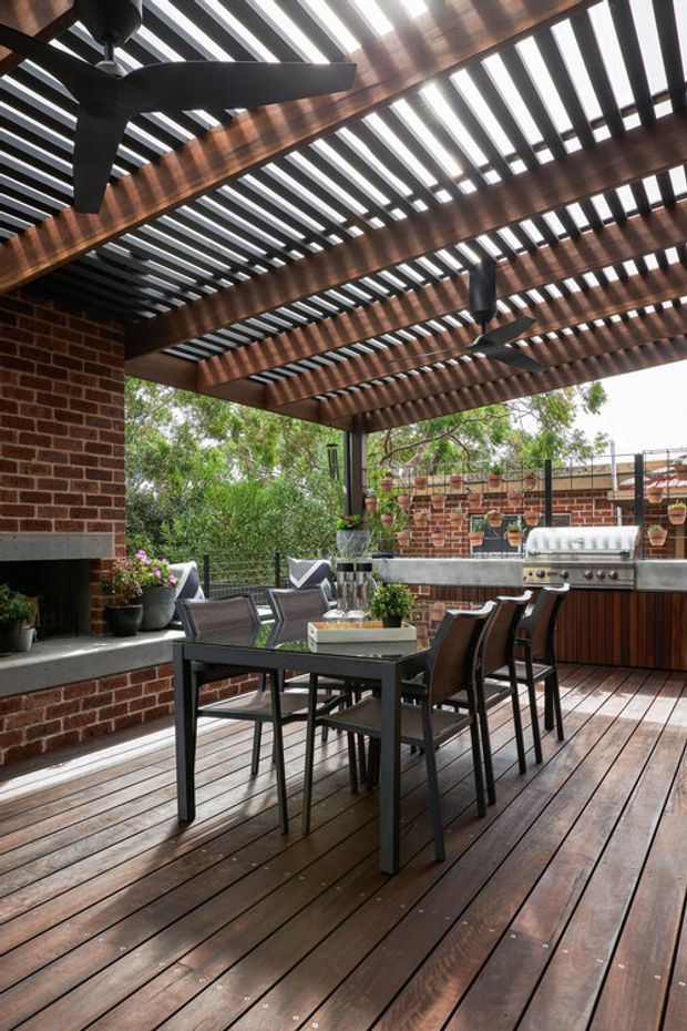 Covered outdoor entertaining terrace with a dark timber deck, exposed brick fireplace and walls, timber pergola with slatted roof, ceiling fans, dining table and chairs, and a built-in outdoor kitchen with barbecue. The space has a warm, sheltered, contemporary-industrial character and overlooks leafy surroundings.