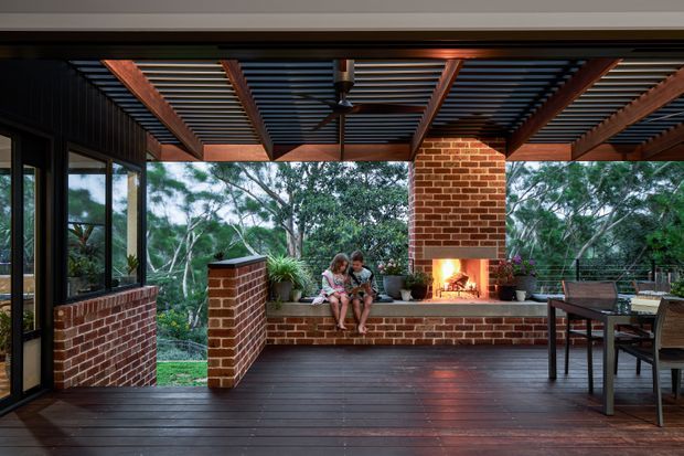 A covered outdoor entertaining terrace surrounded by bushland, featuring dark timber decking, extensive glazing, red brick walls and fireplace, and a timber pergola with dark metal slatted roof. A built-in brick bench and dining table create a warm outdoor living area, with a lit fireplace as the focal point.
