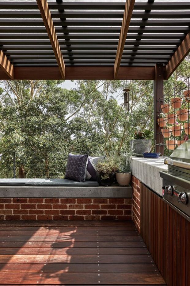 A compact elevated outdoor entertaining deck surrounded by leafy trees. The space features dark timber decking, a red brick built-in bench with a grey concrete cap and cushions, a stainless-steel barbecue with timber-clad cabinetry, potted plants, wire balustrading, and a timber-and-dark-metal pergola overhead. The image has a warm, natural, private atmosphere with strong sunlight and shadows.