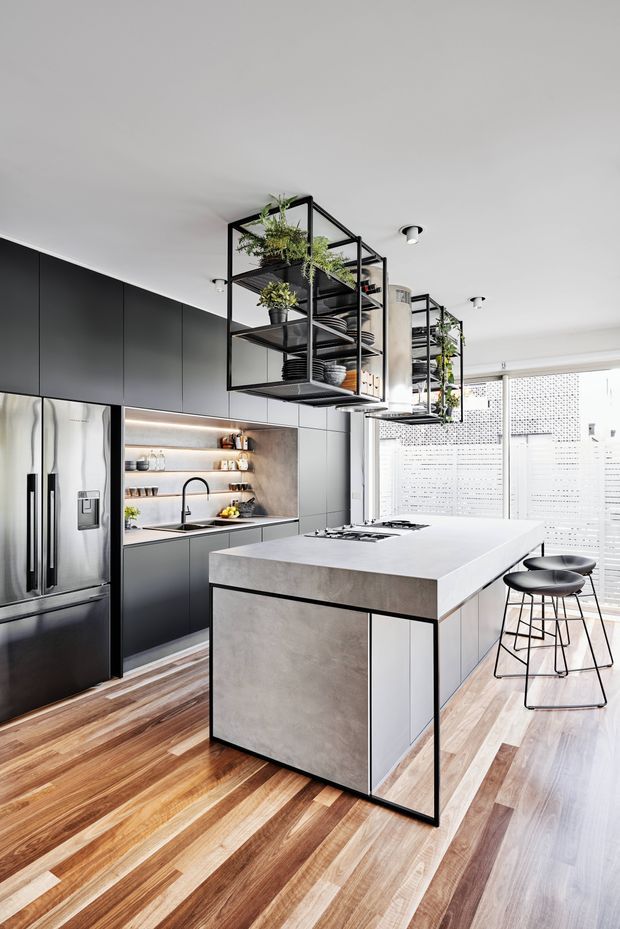 A bright contemporary kitchen featuring matte dark built-in cabinetry, a large pale concrete-look island with integrated cooktop, open black metal shelving suspended from the ceiling, stainless-steel appliances, timber flooring, and a bank of perforated screening and glazing at the rear. Black bar stools and hanging plants add contrast and texture.