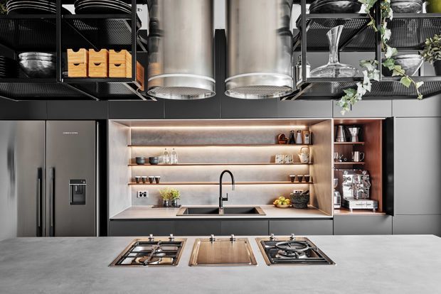 A highly refined contemporary kitchen viewed from the island, featuring a broad grey stone island benchtop with integrated gas cooktops and a central timber chopping board. The back wall has dark cabinetry, a stainless-steel refrigerator, an undermount sink with black tapware, open illuminated timber shelving, and a coffee station. Large cylindrical stainless-steel rangehood ducts and black open shelving overhead create an industrial-inspired focal point. Warm integrated lighting, timber accents, and trailing greenery add contrast to the monochromatic palette.