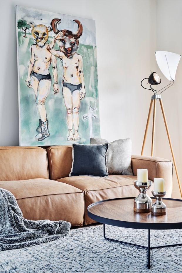 A stylish living room featuring a tan leather sectional sofa with navy and grey cushions, a round dark-edged coffee table with silver candle holders, a textured grey area rug, and a sculptural tripod floor lamp. A large expressive figurative artwork leans against the pale wall behind the sofa, creating a contemporary, eclectic focal point.