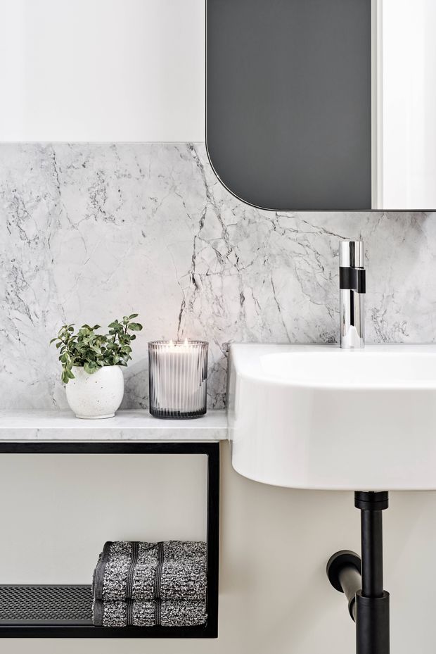 Close-up of a refined contemporary bathroom vanity. A white wall-mounted basin with a chrome and black tap sits against a white-grey veined marble wall finish. A matching marble shelf holds a small plant and ribbed glass candle, while a black-framed open vanity shelf below contains a folded patterned towel. A large rounded-corner dark mirror and black plumbing details add graphic contrast.