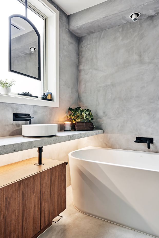 A refined contemporary bathroom featuring a freestanding white bathtub, white vessel basin, matte black wall-mounted tapware, and a floating timber vanity. The walls and benchtop have a raw, mottled grey concrete finish, complemented by a large black-framed mirror, recessed downlights, and subtle indoor greenery.