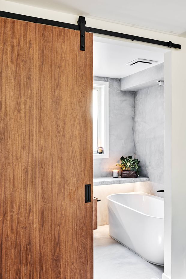 A bright contemporary bathroom viewed through a partially open sliding barn door. The room features a freestanding white bathtub, grey concrete-look wall and floor finishes, a narrow window, recessed ceiling ventilation, and a built-in ledge with a candle and plant. The door is warm timber with black metal barn-door hardware.