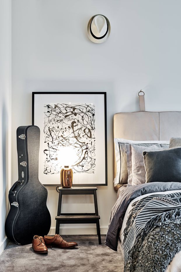 A softly lit, styled bedroom vignette with a neutral upholstered bed, layered grey and cream bedding, a black-framed abstract artwork, a small dark side table with a ceramic lamp, a guitar case, leather shoes, and a sculptural wall light. The room has a calm, sophisticated contemporary feel with warm tactile textures.