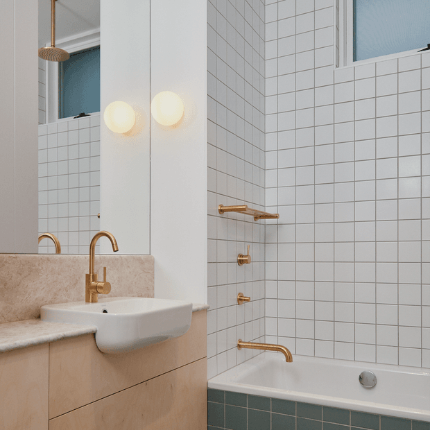 A compact contemporary bathroom with a wall-mounted white basin set into a pale stone vanity, a mirrored cabinet, white square wall tiles, and a bathtub with dark green tiled sides. Brushed brass tapware, shower fittings, and spherical pendant lights add warm metallic accents.