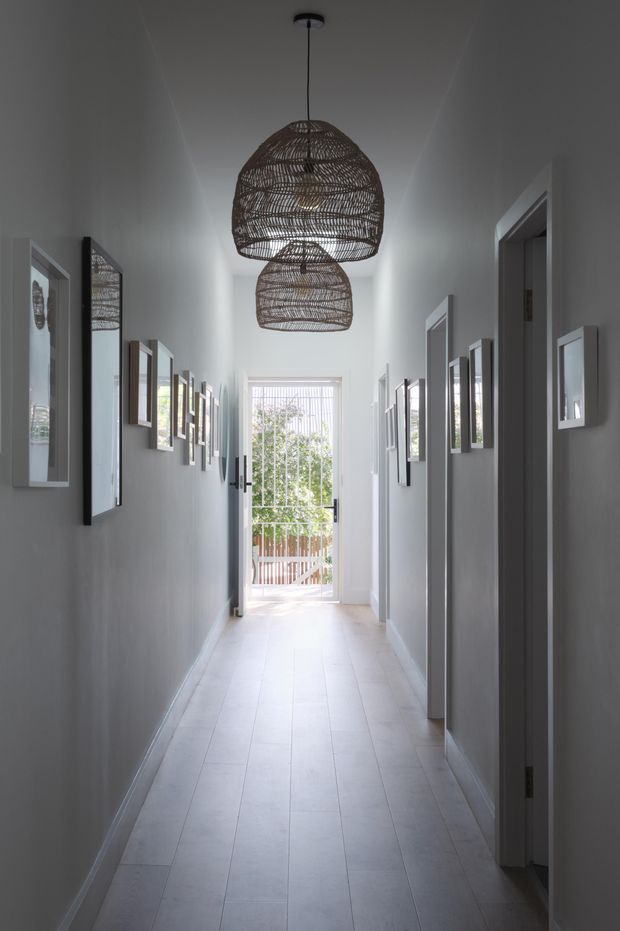 A long, narrow residential hallway with pale grey walls, white trim, and light tiled flooring. Multiple framed artworks and mirrors line both walls, while two large woven pendant lights hang along the ceiling. Glazed doors at the far end open toward a bright outdoor area, creating a calm, airy perspective.