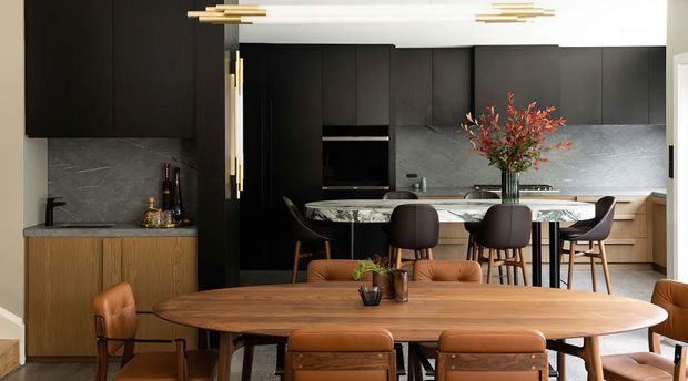 A refined open-plan kitchen and dining area with dark full-height cabinetry, grey stone surfaces, warm timber lower cabinetry, and a large sculptural timber dining table. A marble- or stone-topped island with bar seating sits behind the dining area, complemented by leather dining chairs, brass-accented lighting, and contemporary styling.