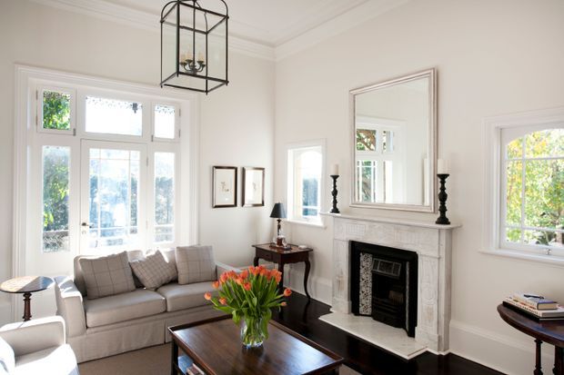 Bright, traditionally styled living room with a pale upholstered sofa and armchair, dark wood coffee and side tables, a white decorative fireplace mantel, large wall mirror, framed artwork, tall multi-pane windows and a black metal lantern pendant. The room has white painted architectural detailing, moulded cornices and trim, with greenery visible outside.