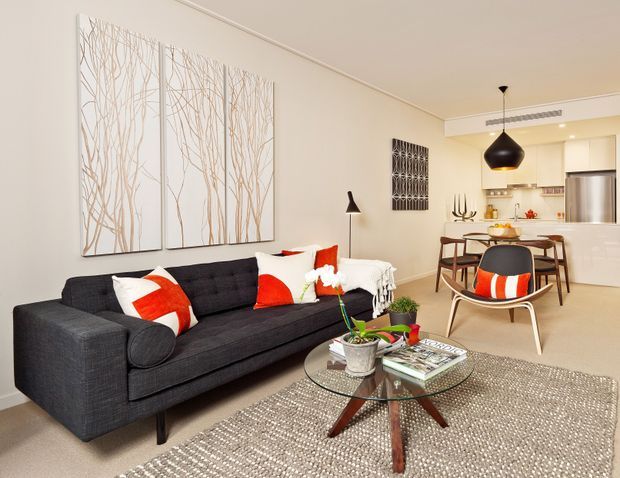 Open-plan living and dining area with a contemporary black sofa, patterned accent cushions, glass coffee table, woven rug, and a compact dining zone leading into a white kitchen. Neutral walls and cabinetry are complemented by black lighting and red-orange accents.