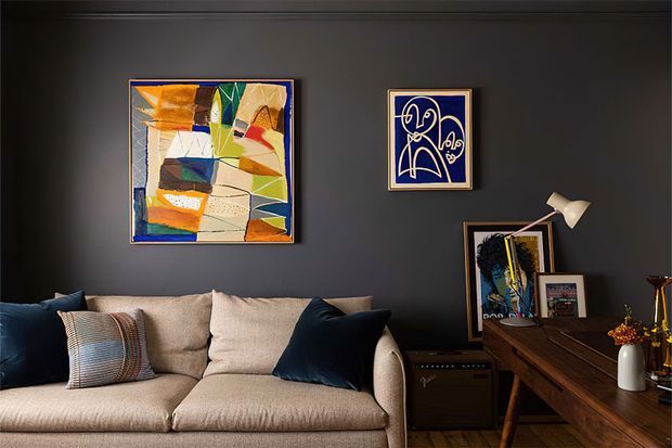 A moody, refined living room with a large beige upholstered sofa, dark navy charcoal walls, blue and patterned cushions, abstract artwork, framed photography, and a timber console or desk styled with a table lamp and decorative objects.