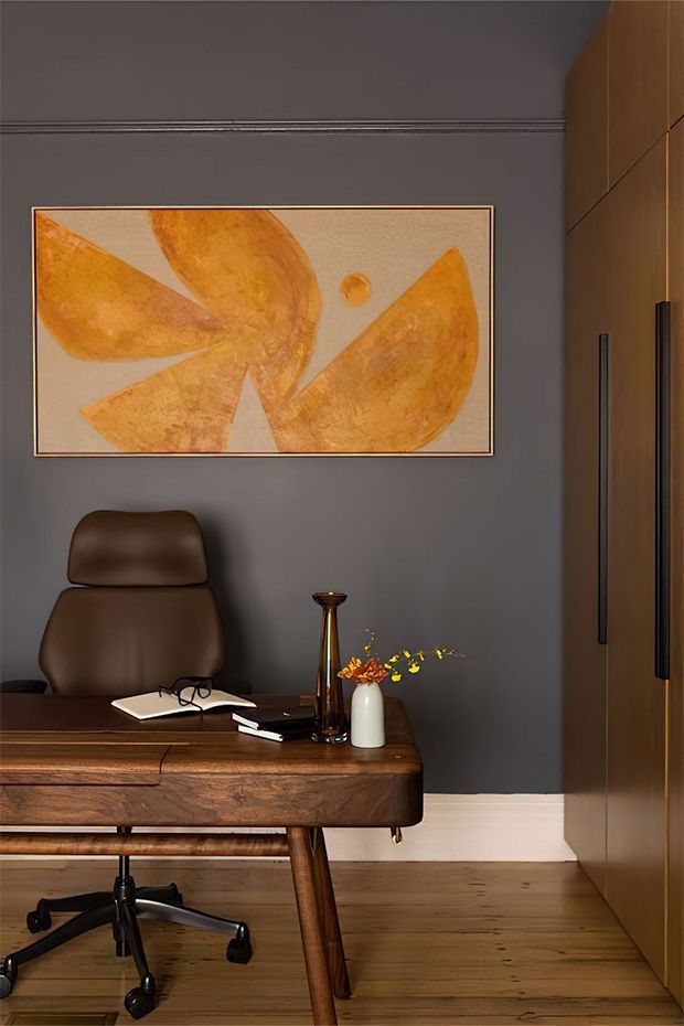 A compact, moody home office with a dark grey painted wall, warm timber flooring, a substantial mid-century-style wooden desk, and a brown leather office chair. A large abstract artwork in ochre and beige hangs above the desk, while full-height timber cabinetry with long dark handles occupies the right side. Small decorative objects and stationery sit on the desk, creating a refined, warm workspace.