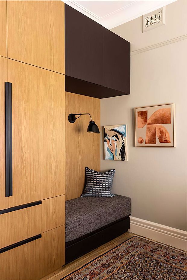 A compact, refined built-in entryway or hallway nook featuring full-height light timber cabinetry with long black pulls, a dark overhead storage volume, and a built-in upholstered bench with patterned cushion. Warm wall lighting, framed abstract artwork, decorative ceiling detailing, and a traditional patterned rug add character to the neutral interior.
