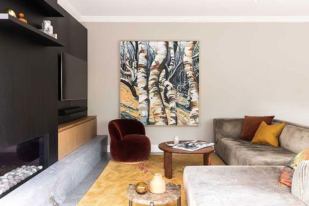 A refined living room with a dark built-in media and fireplace wall, warm timber cabinetry, a pale stone hearth, neutral walls with decorative crown moulding, and a large sectional sofa arranged around a round wooden coffee table. A deep burgundy accent chair, mustard and rust cushions, a patterned rug, and botanical artwork add warmth and colour.