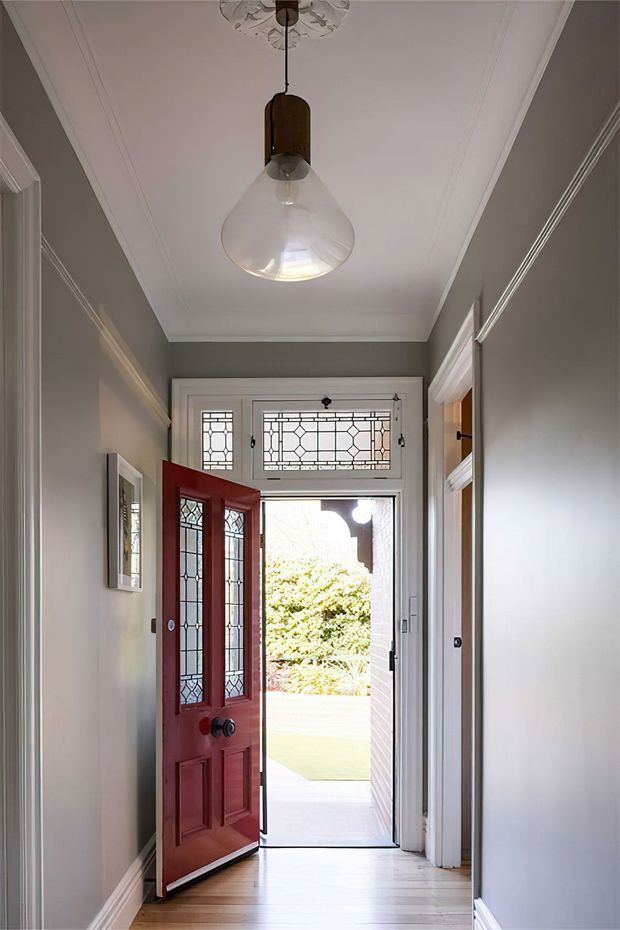 A narrow residential entry hall viewed from inside, featuring grey painted walls, white decorative moulding and trim, pale timber flooring, and a deep red timber front door with leadlight glass panels. A matching leadlight transom and sidelights bring daylight into the hallway, while a pendant light hangs from a decorative ceiling rose. The open doorway reveals a small landscaped exterior beyond.