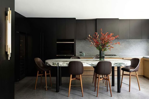 Contemporary kitchen and dining area with dark matte cabinetry, a large veined stone dining table, upholstered timber-leg dining chairs, and a textured grey splashback. A sculptural wall light and floral arrangement add warm accents to the otherwise monochromatic space.