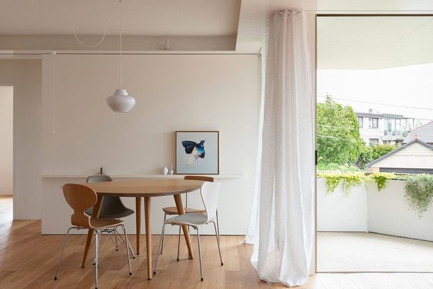 Bright, minimalist dining area with a light timber dining table and mixed mid-century-style chairs, set against a simple white built-in wall with a slim shelf and framed artwork. Full-height sheer curtains open onto a small balcony or terrace, with warm timber flooring and abundant natural light.