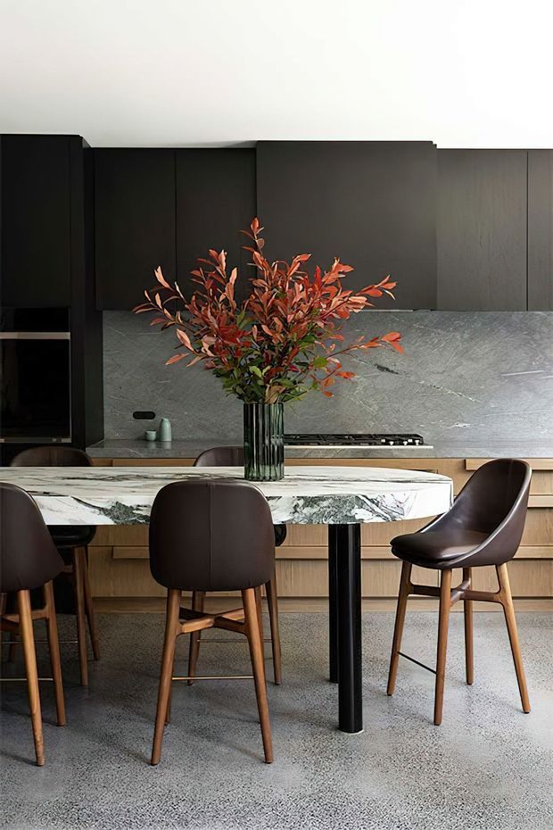 A sophisticated contemporary kitchen and dining area with dark matte cabinetry, a large grey veined stone splashback, timber lower cabinetry, and a marble-topped dining table with black pedestal legs. Dark brown leather dining chairs with timber legs surround the table, while a tall glass vase with red foliage forms a central accent. The space has a restrained, warm neutral palette and polished yet inviting styling.