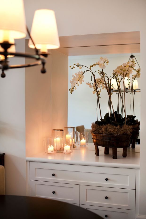 A warmly lit interior vignette featuring a white paneled chest of drawers beneath a large wall mirror. Multiple candles in glass holders and a large potted orchid arrangement decorate the surface, while a dark multi-arm pendant light is reflected in the mirror. The room has a calm, refined, softly traditional appearance.