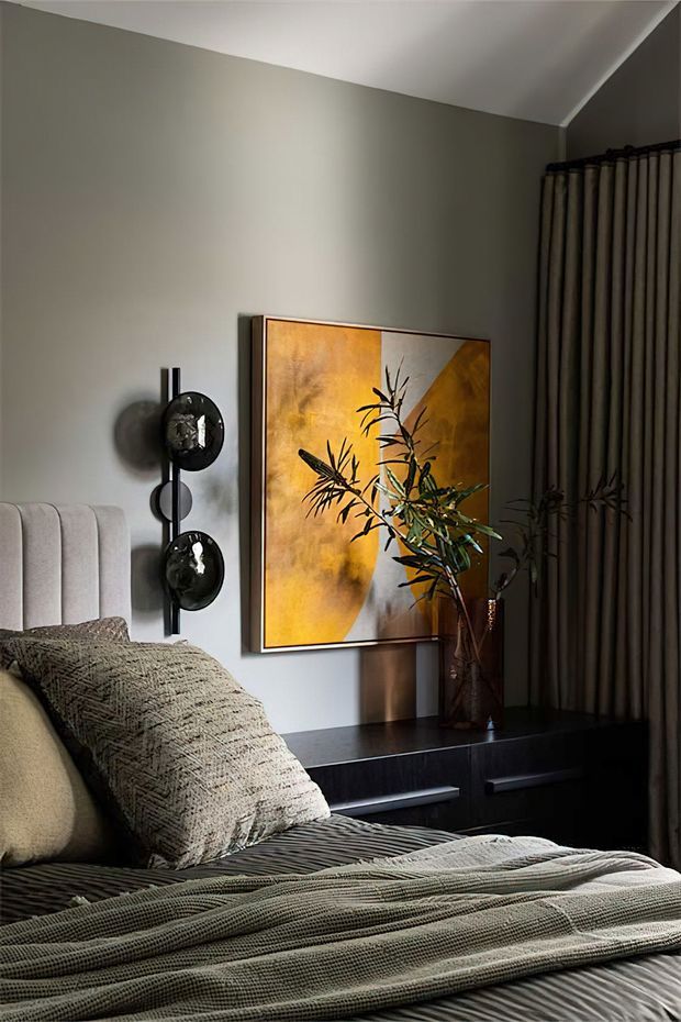 A moody, refined bedroom interior featuring a channel-tufted upholstered bedhead, layered textured bedding, a dark built-in bedside cabinet, striped floor-to-ceiling curtains, and a large warm-toned abstract artwork. Sculptural black wall lighting and a tall decorative branch arrangement add dramatic accents.