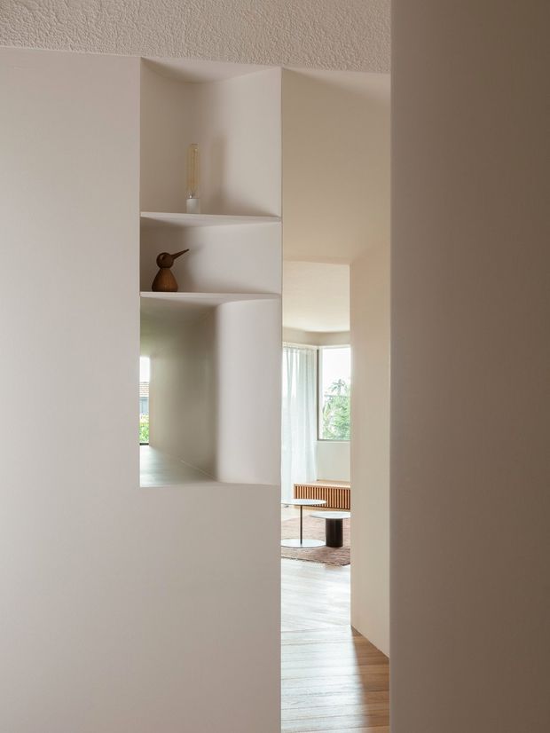 A minimalist interior view through a narrow opening in a white built-in partition wall. The partition incorporates recessed open display niches with a small vase and decorative object. Beyond it, a bright living room is partially visible with floor-to-ceiling curtains, a large window, timber flooring, a low coffee table, and a slatted timber console or bench. The space has soft natural light, warm neutral finishes, and clean sculptural wall geometry.