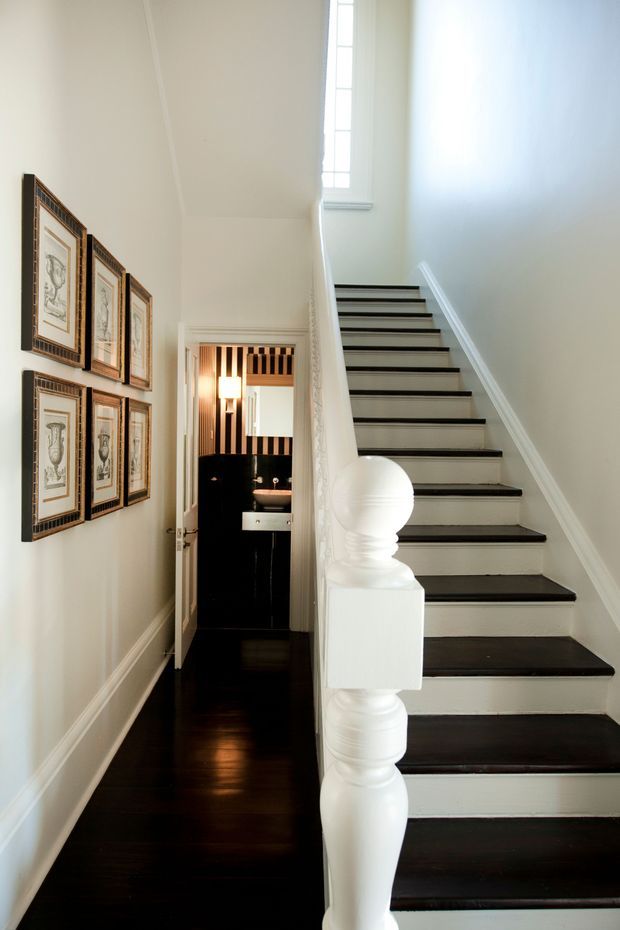 A narrow, elegant interior hallway with a dark timber floor and a striking staircase. The staircase has dark treads, white risers, ornate white turned balusters and a sculpted newel post, leading toward a tall narrow window. Framed artwork is arranged along the left wall, and an open doorway reveals a small dark bathroom or powder room with patterned wall detailing and a vanity. The overall presentation is refined and traditional with strong black-and-white contrast.