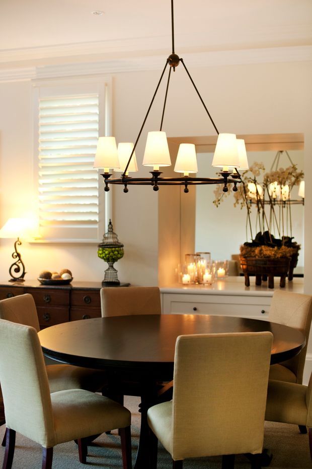 A warm, refined dining room featuring a large round dark wood dining table surrounded by upholstered dining chairs. A prominent black metal chandelier with multiple cream fabric shades hangs above the table. The room has light painted walls with decorative moulding, a white plantation-shutter window, dark wood sideboard cabinetry, a built-in white display cabinet, and a large mirror reflecting floral decor and candlelight.