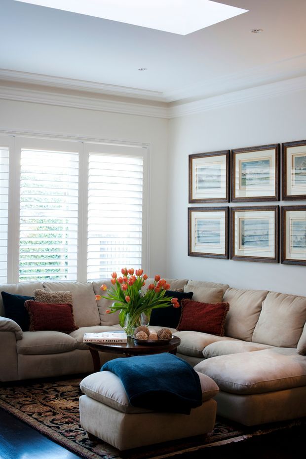 A bright, refined living room with a large cream sectional sofa, upholstered ottoman, dark wood coffee table, patterned area rug, framed artwork, white plantation shutters, and ornate crown moulding. A large rectangular skylight adds natural light, while blue and deep red accents provide contrast.