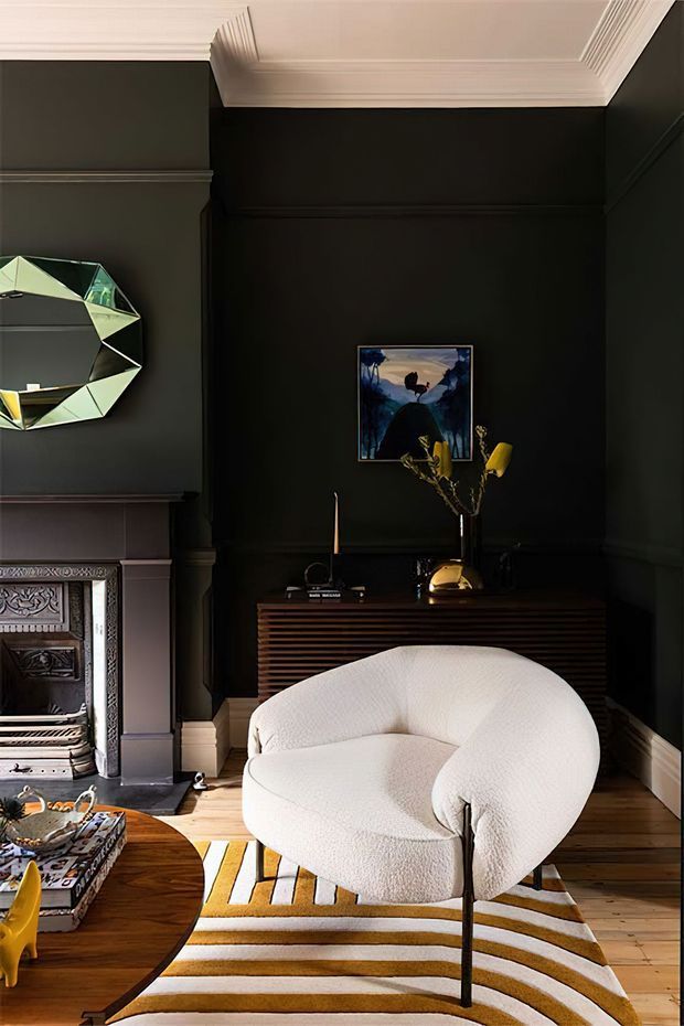 A richly styled living room with deep charcoal-black walls, ornate white crown moulding and wall panelling, and a traditional fireplace with decorative metalwork. A sculptural cream boucle lounge chair sits on a bold curved-stripe rug, with a dark timber slatted console, artwork, sculptural mirror, and coffee table completing the moody, eclectic interior.