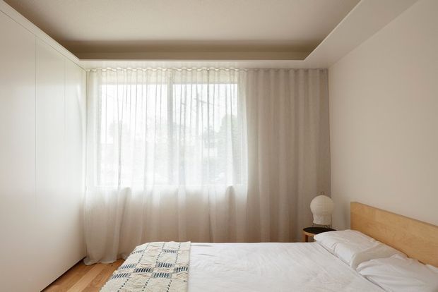 A serene, minimalist bedroom with a low-profile bed, pale timber headboard, white bedding, sheer floor-to-ceiling curtains, integrated white wall storage, and a recessed ceiling detail. Natural light creates a soft, calm atmosphere.