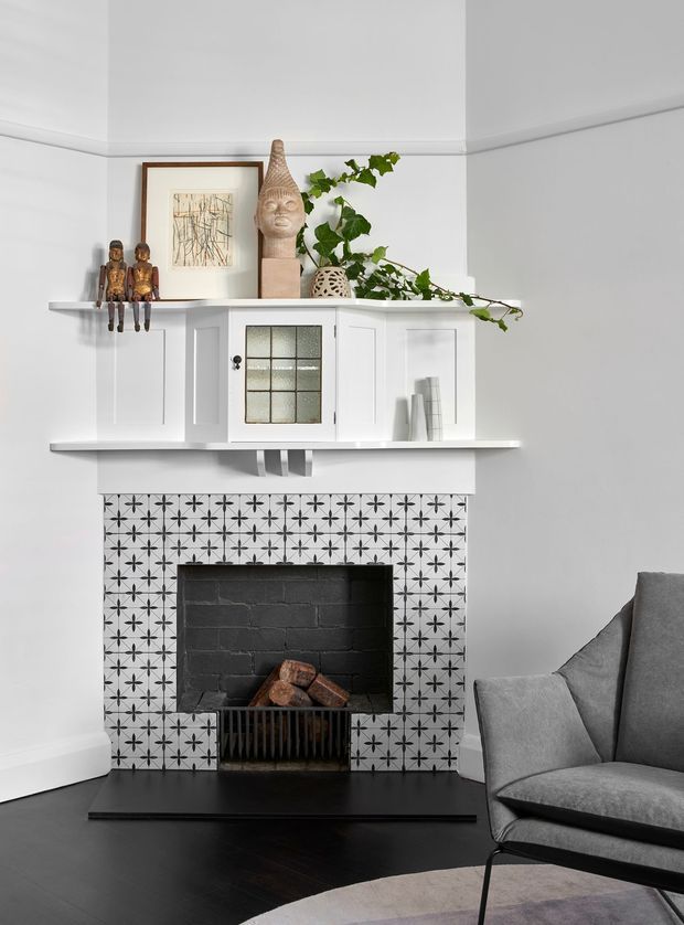 A refined living room corner centered on a traditional fireplace with a bold black-and-white geometric tiled surround, dark hearth, and black-painted firebox. White built-in shelving and cabinetry sit above the mantel, displaying artwork, carved figures, ceramics, books, and trailing greenery. A grey upholstered armchair occupies the foreground, with dark timber flooring and a neutral patterned rug completing the scene.