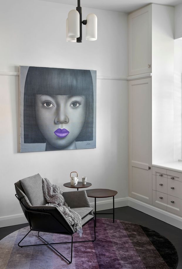 A refined sitting nook with a black metal lounge chair, layered grey throws, and two dark wood nesting side tables. The room features white paneled walls, full-height built-in cabinetry, a bright window seat or low cabinetry beneath the window, a sculptural black pendant light, and a large monochromatic portrait artwork with purple lips. A muted purple and charcoal area rug adds colour and texture.