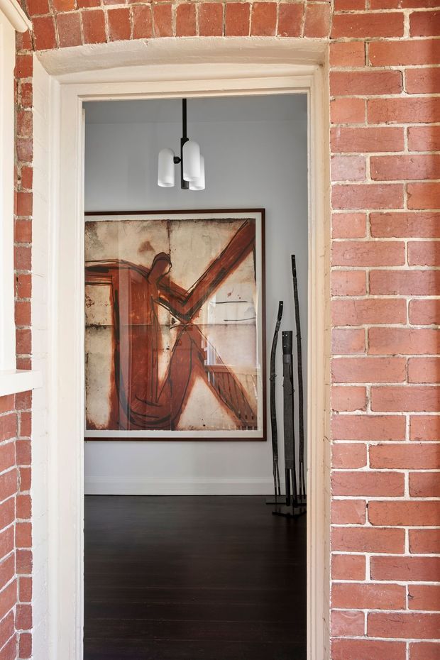View through a white-trimmed interior doorway in a red brick building, looking into a refined room with dark timber flooring, pale walls, a large abstract artwork, and a modern white pendant light. A black sculptural stand or decorative object is positioned near the artwork. The image emphasizes the brick surround, doorway detailing, and the transition into the adjacent interior.
