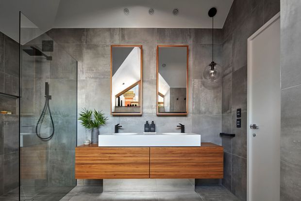 Contemporary bathroom with large grey concrete-look wall and floor tiles, a frameless glass shower, floating timber double vanity with a white basin, twin mirrors, black tapware and wall-mounted fixtures. A partially visible pitched ceiling and warm timber elements are reflected in the mirrors.