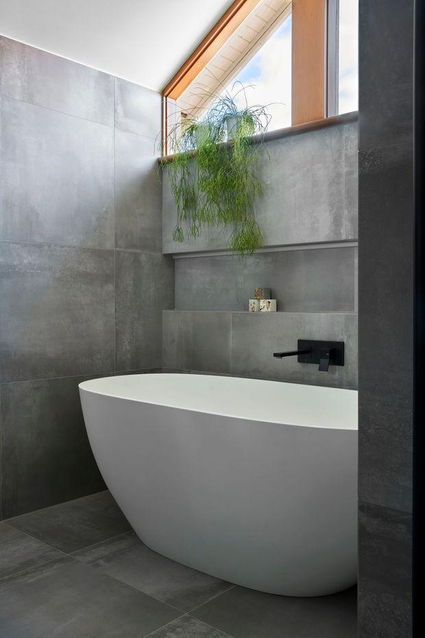 A contemporary bathroom featuring a freestanding white bathtub set against large grey concrete-look wall and floor tiles. A black wall-mounted tap and recessed shelf provide contrast, while a timber-framed skylight or high window brings in natural light above a cascading green plant.