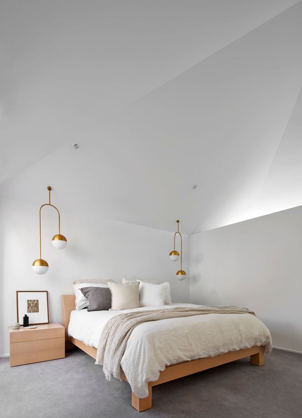 A serene contemporary bedroom with a low timber bed, white bedding, layered neutral throws and pillows, and a simple timber bedside table. The room has pale grey walls, grey carpet, a sharply angled white ceiling, and distinctive suspended brass-and-glass pendant lights.