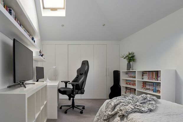 Bright, compact contemporary bedroom and gaming/reading space with built-in white cabinetry, a large television, gaming chair, desk, bookshelf filled with books, skylight, and a bed in the foreground. The room uses a restrained white and grey palette with soft natural light.