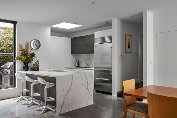 Contemporary open-plan kitchen and dining area with a large white marble-look island, waterfall-style veined surface, white cabinetry, dark wood overhead cabinetry, stainless-steel appliances, and three white bar stools. A timber dining table and chairs occupy the foreground, with a large window and skylight bringing in natural light.