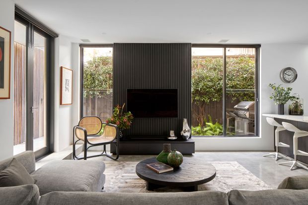A bright, contemporary living room with a large sectional sofa, round coffee table, patterned rug, lounge chair, and wall-mounted television set into a dark vertical-ribbed feature wall. Full-height glazing and a glazed door open to a planted courtyard, while a compact bar or kitchen counter with stools is visible to the right.