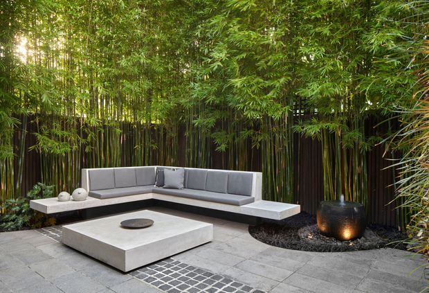 A contemporary outdoor lounge courtyard surrounded by dense bamboo screening. A built-in L-shaped grey sectional sits around a low square concrete coffee table and circular fire feature, with large dark planters and grey tiled paving creating a private, serene setting.