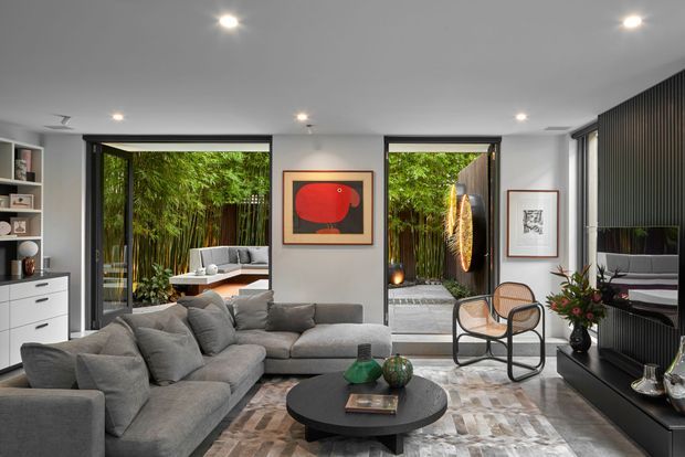 A contemporary living room with a large grey sectional sofa, round black coffee table, built-in shelving and cabinetry, and framed artwork. Full-height glazed doors open toward a lush planted courtyard, while a dark vertical-slat feature wall and recessed ceiling lighting add contrast. The room has a refined, calm, indoor-outdoor character.