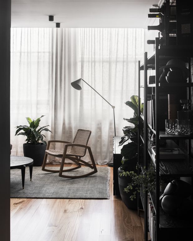 A softly lit contemporary living room with sheer floor-to-ceiling curtains, pale timber flooring, a textured grey rug, a woven wooden rocking chair, a small dark side table, an arched floor lamp, abundant potted greenery, and a tall black open shelving unit filled with books, objects, glassware and decor.